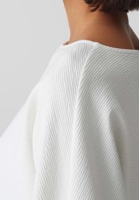 Haut blanc texturé avec un motif côtelé, doté d'un large décolleté et d'une coupe drapée, mettant en valeur un tissu doux et des détails subtils.