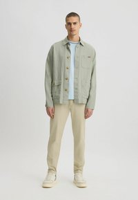 Giacca verde chiaro con bottoni, tasca sul petto e due tasche laterali, indossata sopra una camicia azzurra chiara e pantaloni beige con sneakers.