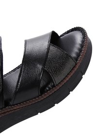 Black leather sandal featuring cross-straps, a textured surface, padded lining, and a thick black sole with a brown braided accent.