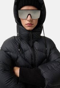 Sort puffervest med hætte, der har et quiltet design og høj krave. Modellen bærer store refleksive solbriller og en sort strikket hue.