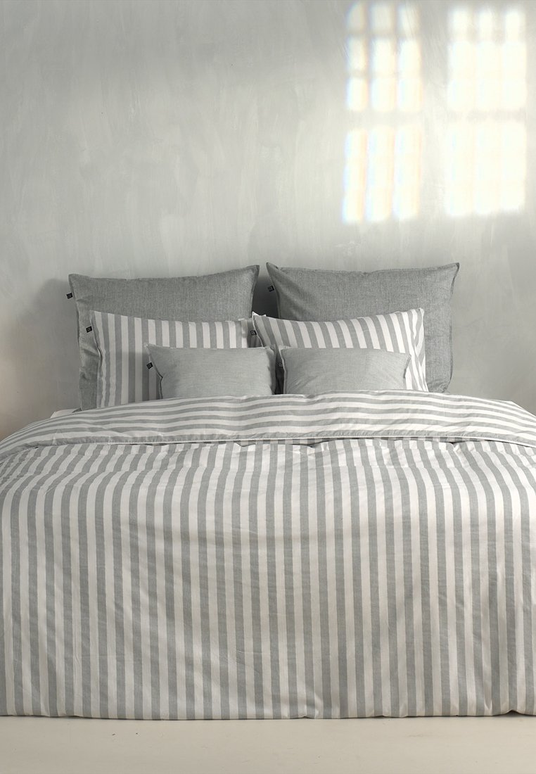 Set di biancheria da letto con copriletto a righe grigie e bianche e cuscini in un morbido tessuto strutturato, sistemati su un letto di colore chiaro.