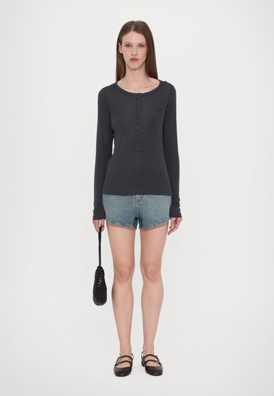 Camisa de manga larga azul marino con botonera, combinada con pantalones cortos de denim. La modelo sostiene un pequeño bolso negro y lleva unos zapatos marrones.