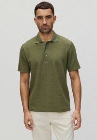 Oliwkowa koszulka polo o teksturowanym wykończeniu, krótki rękaw, zapięcie na trzy guziki i prążkowany kołnierzyk. Noszona z jasnymi spodniami.