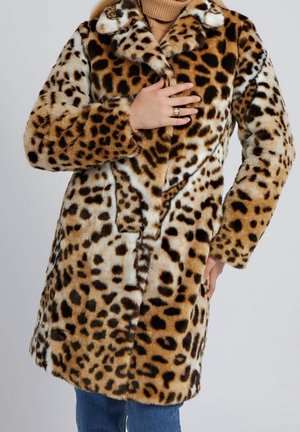 Leopardmønstret faux fur-frakke over beige rullekrave, båret med blå jeans, hånd hvilende på bryst og hofte, guld ringe på fingre.