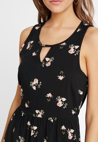 Kvinna i en ärmlös svart klänning med små rosa och vita blommor och en skålad halsringning, visad från axlarna till midjan.