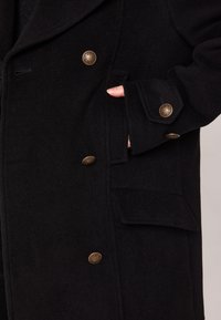 Cappotto di lana nera con design doppiopetto, bottoni decorativi dorati e tasche laterali con finitura testurizzata.