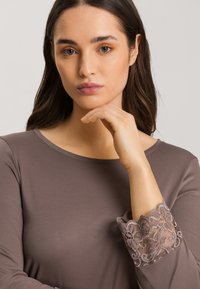 Mujer con cabello largo y oscuro, vistiendo una blusa en tono malva con puños de encaje, apoyando su barbilla en su mano frente a un fondo sencillo.