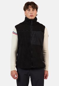 Gilet in pile nero con colletto alto, zip frontale e tasca sul petto con zip. Presenta un pannello nero liscio e una texture a contrasto.