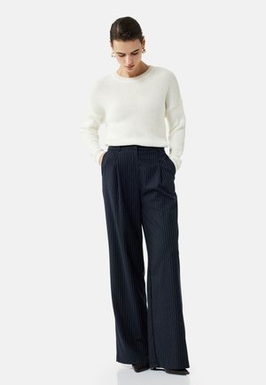 Suéter blanco de punto con relieve combinado con pantalones de pierna ancha a rayas marinas. El modelo tiene las manos en los bolsillos, de pie sobre zapatos oscuros.