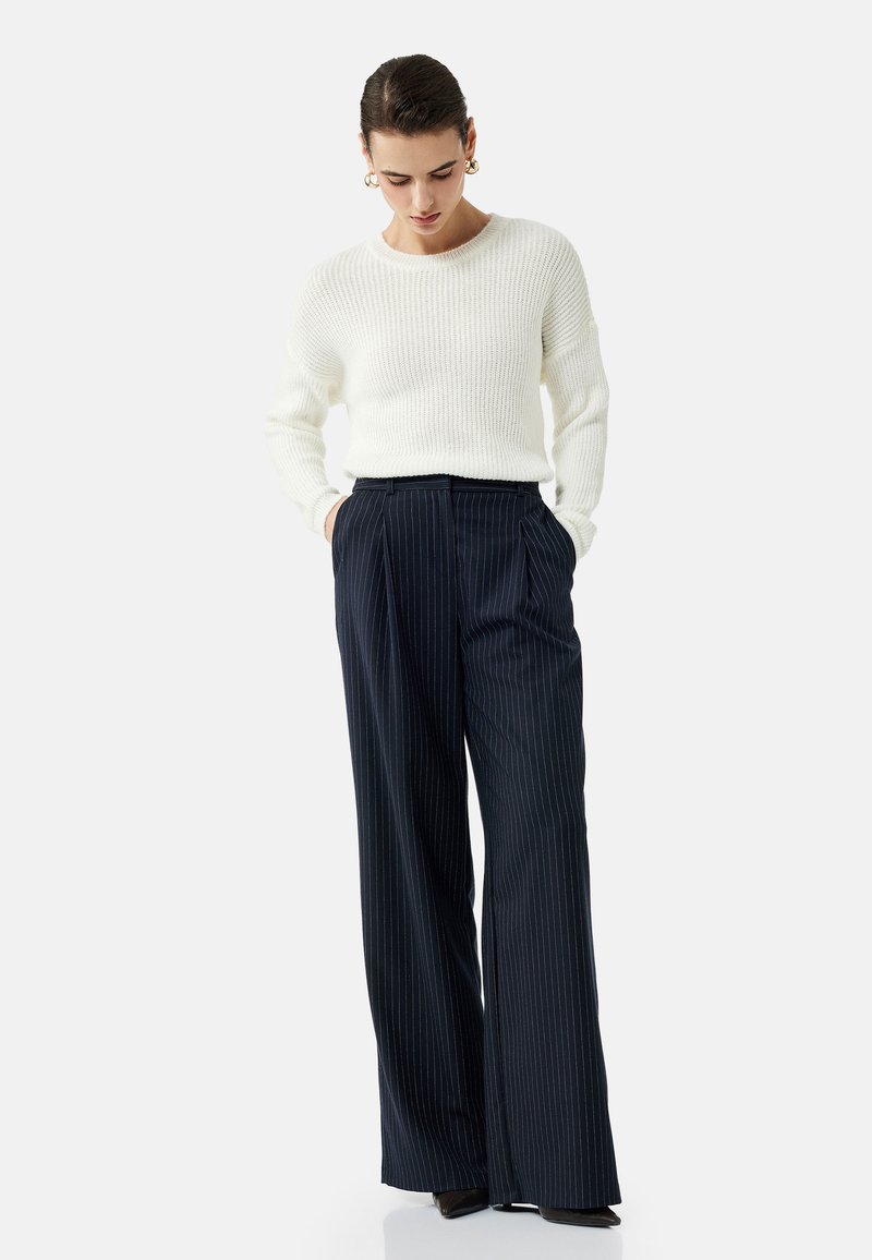 Witte geribbelde gebreide trui gecombineerd met marineblauwe pinstripe wijde broek. Het model heeft de handen in de zakken en staat op donkere schoenen.