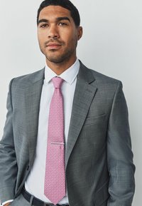 Grauer strukturiertes Sakko über einem weißen Hemd, mit einer rosa gemusterten Krawatte und einer silbernen Krawattennadel. Sauberes und professionelles Erscheinungsbild.
