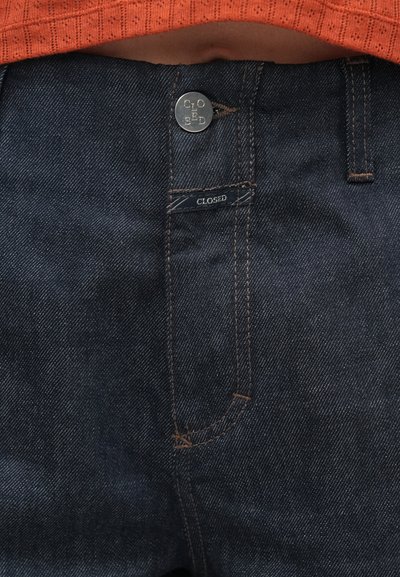 Σκούρο τζιν παντελόνι με κουμπιά κλεισίματος, εμφανή ραφές και ετικέτα με τη λέξη "CLOSED". Διαθέτει ίσια γραμμή και τυπικό σχέδιο τσέπης.