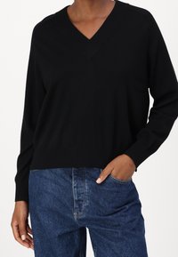 Pull en tricot noir avec un décolleté en V et un ourlet côtelé. Présente des manches longues et une coupe décontractée, associé à un jean en denim bleu foncé.