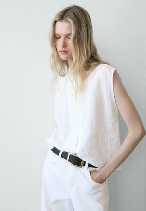 Donna bionda con capelli ondulati indossa una camicetta bianca senza maniche, pantaloni bianchi e cintura nera con fibbia d'argento, in piedi davanti a una parete chiara e uniforme.