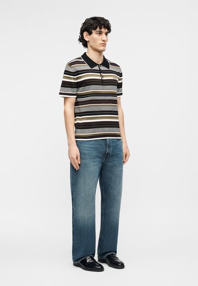 Άνδρας με κοντά σγουρά μαλλιά, που φοράει ριγέ κοντομάνικο πόλο, μπλε τζιν και μαύρα παπούτσια, στέκεται μπροστά σε απλό φόντο.