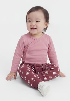 Tout-petit souriant assis les jambes croisées, portant un haut à manches longues rose, un pantalon à fleurs et des chaussures blanches sur fond blanc.