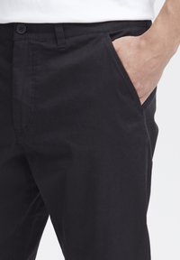 Pantalonii negri din bumbac au o croială ajustată, două buzunare laterale și o închidere cu un singur nasture, având o textură netedă și uniformă.