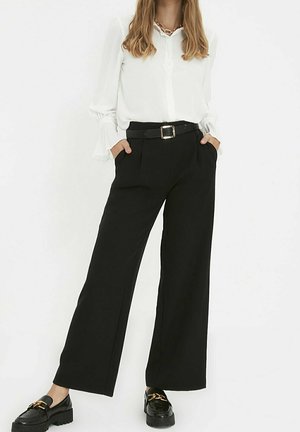 Frau trägt eine weiße Langarmbluse, die in eine hochtaillierte, weite schwarze Hose mit schwarzem Gürtel und schwarzen Loafers mit goldener Kettendetail gesteckt ist.