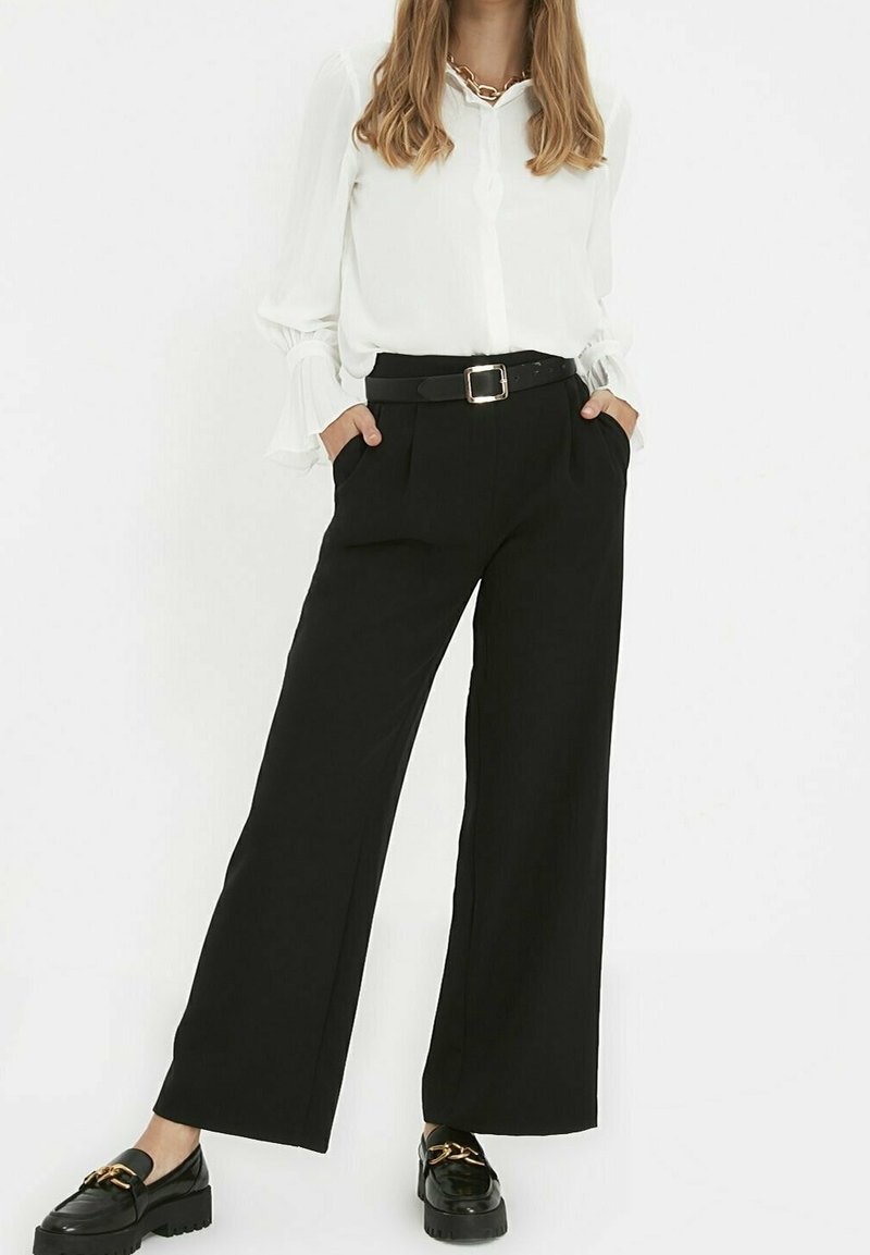 Femme portant un chemisier blanc à manches longues rentré dans un pantalon large noir taille haute, avec une ceinture noire et des mocassins noirs ornés d'une chaîne dorée.