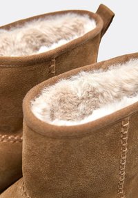 Brązowe botki z zamszu z miękkim beżowym futerkiem w środku, przeszyciami i pętelką z tyłu ułatwiającą zakładanie. Teksturowana powierzchnia zewnętrzna.