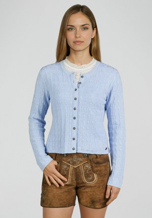 Vrouw die een lichtblauw vest met knopen draagt over een wit kanten topje en traditionele bruine lederen korte broek met borduursels, staand met één hand in de zak.