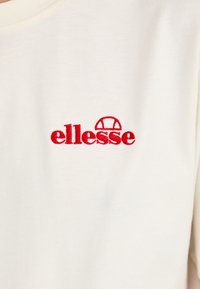 T-shirt en coton blanc avec un logo "ellesse" brodé en rouge. Le tissu a une texture lisse avec un design classique à col rond.