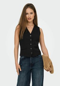 Junge Frau mit langen braunen Haaren trägt eine schwarze geknöpfte Weste, dunkelblaue Jeans und hält eine beige geflochtene Tasche in der rechten Hand vor einem schlichten Hintergrund.