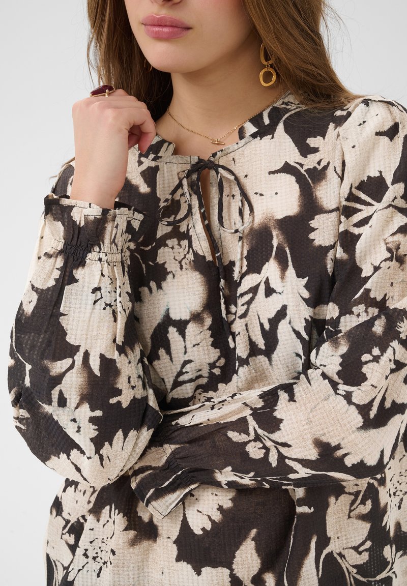 Femme portant un chemisier beige et noir à motifs floraux avec une encolure en forme de goutte d'eau nouée avec un cordon, un collier en or, une bague et des boucles d'oreilles circulaires en or.