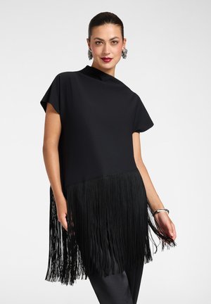 Femme portant une tunique noire à franges avec des manches courtes, des boucles d'oreilles argentées en forme d’anneau et un bracelet argenté, debout devant un fond uni.