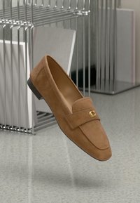 Mocassini in suede marrone con suola piatta, caratterizzati da un accento con logo dorato e una punta cucita. Sullo sfondo ci sono scaffali in metallo.