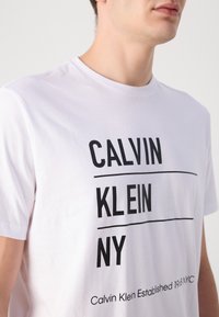 Bijela pamučna majica s odvažnim crnim natpisima: "CALVIN", "KLEIN" i "NY." Sadrži slogan na dnu. Klasičan dizajn s okruglim izrezom.