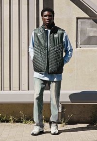 Mørkegrøn quilted vest over en lyseblå langærmet skjorte, parret med lyse denim jeans og hvide sneakers. Står op ad en struktureret væg.