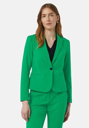 Blazer vert vif en tissu lisse, avec une fermeture à bouton unique, des revers crantés et une silhouette ajustée. Associé à un top noir.
