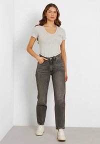 Gray v-neck t-shirt with a small logo, paired with high-waisted faded black jeans. Model wears white sneakers; minimalist background.