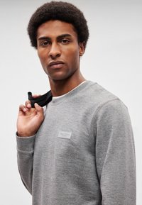 Sweatshirt cinzenta com decote redondo, tecido texturado e uma pequena etiqueta de marca no peito. Modelo segura a gola com um objeto preto.
