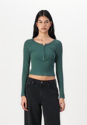 Langärmliges, kurzes Oberteil aus dunkelgrünem, geripptem Stoff mit rundem Halsausschnitt und Knopfverschluss. Kombiniert mit hochgeschnittenen schwarzen Jeans.