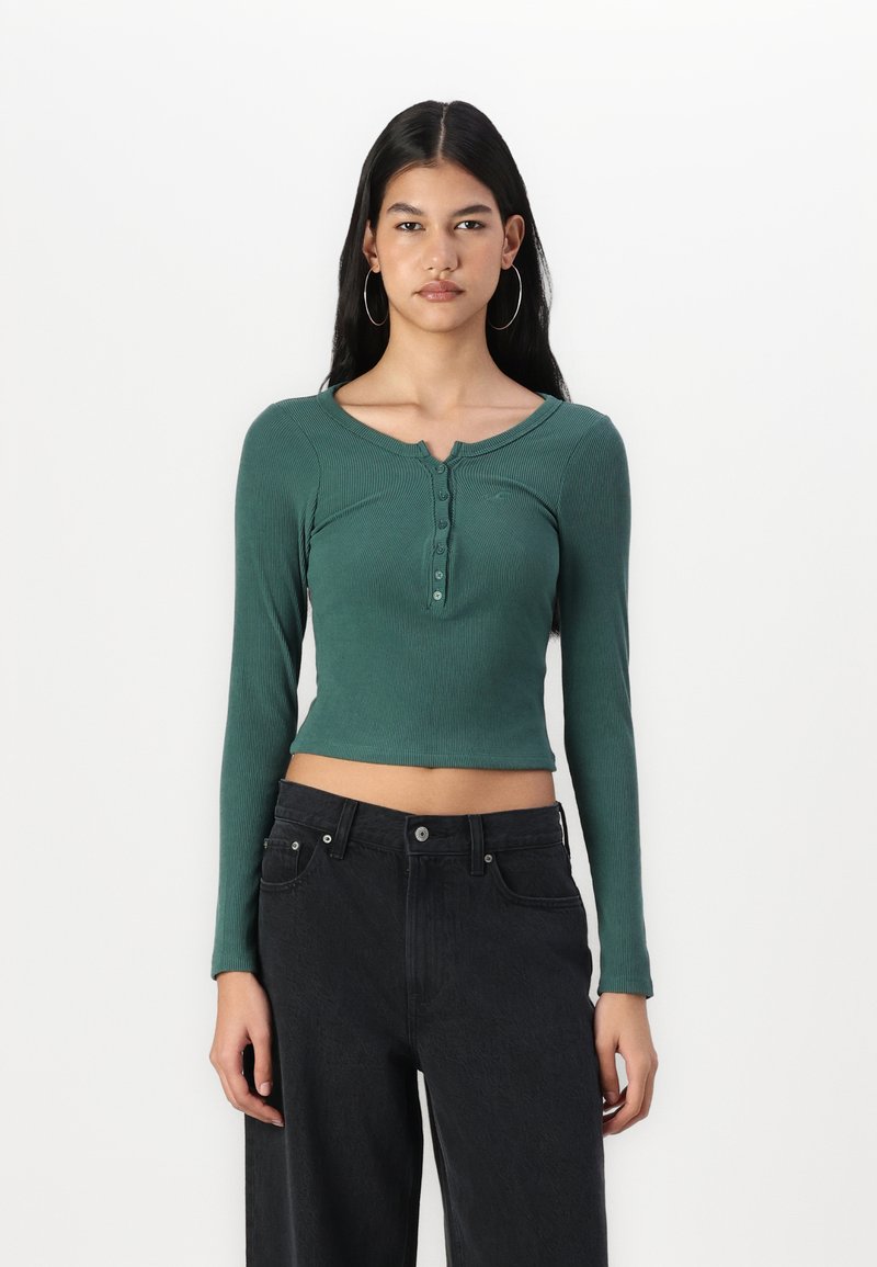 Langermet, cropped topp i mørkegrønn ribbestrikket stoff med rund hals og knappelukking. Kombinert med høyhåndte svarte jeans.
