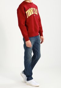 Sudadera roja con "TOMMY JEAN" en amarillo, combinada con jeans azules y zapatillas blancas. La tela parece suave con un corte relajado.