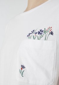 Bílé bavlněné tričko s kapsou na hrudi, zdobené vícebarevn�ým květinovým vyšíváním na kapse a v dolním rohu.
