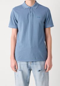 Jasnoniebieska koszulka polo z bawełny, z kołnierzykiem, listwą na dwa guziki i logo na piersi. Połączona z przetartymi dżinsami.