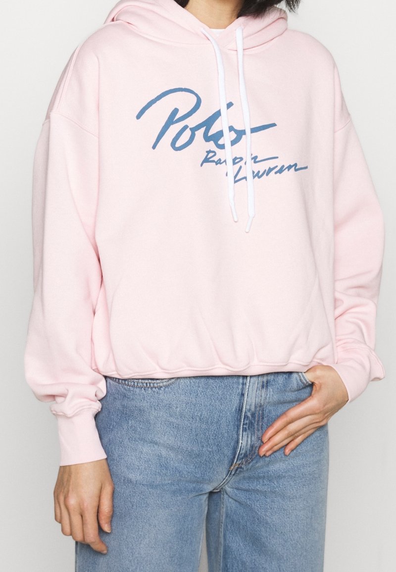 Rosa huvtröja med vita dragsnoddar, med en blå logga "Polo Ralph Lauren" över framsidan. Tyget verkar mjukt och har en avslappnad passform.