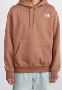 Sudadera marrón con bolsillo canguro, capucha ajustable con cordones y logo blanco en la parte superior izquierda del pecho. Tejido de mezcla de algodón.
