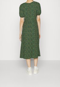Robe midi verte à fleurs avec manches bouffantes, taille cintrée et jupe évasée. Associée à des baskets blanches. Fond uni.