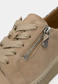 Αθλητικό παπούτσι από μπεζ σουέτ με πλεκτά κορδόνια, πλαϊνό φερμουάρ, ασημένια μεταλλικά στοιχεία και λαβιδωτή ελαστική σόλα. Σημαντικές λεπτομέρειες περιλαμβάνουν μια μικρή δερμάτινη ετικέτα.