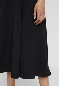 Svart klänning gjord av texturerad tyg, med en flödande design och en asymmetrisk fåll. En hand är försiktigt placerad på tyget.