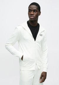 Bela jopa z zadrgo, z zategovalnim kapuco, sprednjimi žepi in majhnim črnim logotipom. Gladek material s klasičnim krojem, kombiniran z ujemajočimi hlačami.