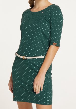 Femme portant une robe verte à pois blancs, longueur genou, avec une fine ceinture blanche, debout avec le bras gauche détendu le long du corps.