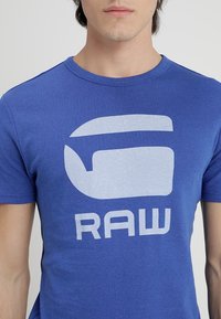 Młody mężczyzna ubrany w niebieską koszulkę z dużą, jasnoniebieską stylizowaną grafiką i napisem "RAW" na piersi.