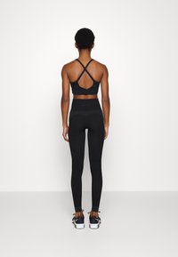 Sostén deportivo negro de diseño cruzado en la espalda; leggings negros de cintura alta con panel texturizado cerca de la cinturilla. Zapatillas negras con suelas blancas.