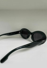 Occhiali da sole ovali neri con lenti scure, dotati di una montatura in plastica lucida e liscia, con curve allungate sulle aste, marchiati con "KOMONO."